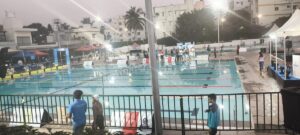 Outdoor swimming competition venue at dusk, showing a floodlit Olympic-size pool with lane markings, event signage, officials, and spectators gathered around the deck.